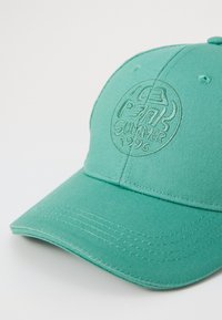 Zelena pamučna kapa s okruglim vizir, s utisnutim logotipom koji glasi "P7K Ljeto 1996." Vidljiva teksturirana tkanina i šivani detalji.