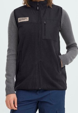 Person iført en sort fleecevest med lynlås på brystlommen og et mærke med teksten "Designed with Dedication" over en grå strikket sweater.
