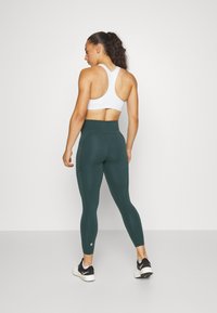 Baltas sportinis liemenukas ir tamsiai žali aukštai juosmeniai leggingai su tekstūruotu paviršiumi. Avalynė juoda su balta padu, su logotipu šone.