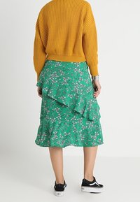 Person wearing a mustard yellow knit sweater, green floral ruffled midi skirt, black and white sneakers, standing against a plain white background.