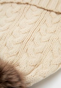 Krémová čepice s copánkovým vzorem, vyrobená z husté texturované příze, s hnědým kožíškovým bambulí na vrcholu.
