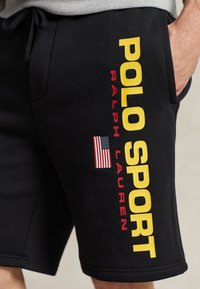 Schwarze Baumwollshorts mit großer gelber "POLO SPORT"-Aufschrift und rotem "RALPH LAUREN", ergänzt durch ein kleines amerikanisches Flaggenakzent.
