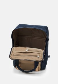 Mochila marina con interior beige. Cuenta con un bolsillo frontal con cremallera y asas resistentes. Material textil suave, forma rectangular y un interior espacioso.