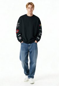 Mladý muž v čiernej mikine s farebnými grafikami na rukávoch, vo voľných modrých džínsach a bielych teniskách, stojí so založenými rukami v vreckách.