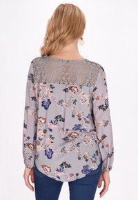 Camisa floral de manga larga en gris claro, con un panel de encaje en la parte posterior y patrones florales multicolores. Detalle de dobladillo curvado.