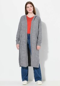 Grå lång cardigan med huva, texturerad tyg, öppen front, sidofickor, bärs över en röd topp och blå jeans, matchad med vita sneakers.