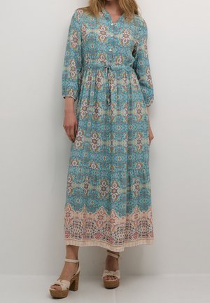 Kvinde iført en langærmet, knap-op turkis maxikjole med blomstrede og geometriske mønstre, kombineret med beige platform-sandaler med høje hæle.