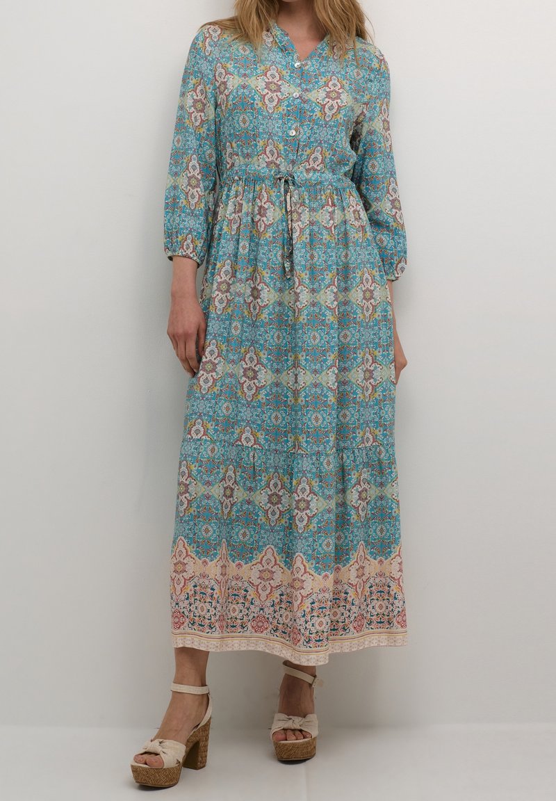 Femme portant une robe maxi boutonnée à manches longues turquoise avec des motifs floraux et géométriques, accompagnée de sandales à talons hauts beiges avec plateforme.