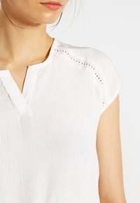 Blusa bianca con tessuto testurizzato, maniche a cappuccio e scollo a V. Delicati dettagli in pizzo lungo le cuciture delle spalle.