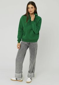 Hoodie en coton vert avec une poche avant, cordon de serrage ajustable et poignets côtelés, associé à un jean en denim gris taille haute et des baskets blanches.
