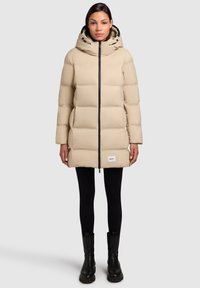 Beige padded puffer coat with a hood, featuring a full front zipper, side pockets, and a brand label at the hem, paired with black leggings and boots.