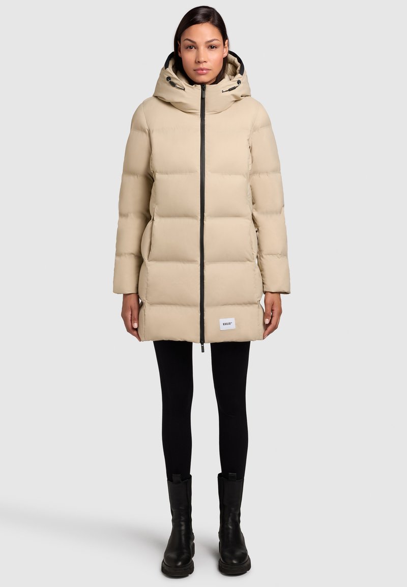 Beige padded puffer coat with a hood, featuring a full front zipper, side pockets, and a brand label at the hem, paired with black leggings and boots.