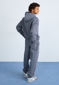 Szara bluza z kapturem i dopasowane dresy, z bocznymi kieszeniami. Materiał wydaje się miękki z lekko teksturowanym wykończeniem. Srebrne sneakersy dopełniają wygląd.