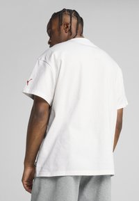Hvid bomulds t-shirt med korte ærmer, der har et rødt logo på venstre skulder. Designet er løst og har en rund halsudskæring.