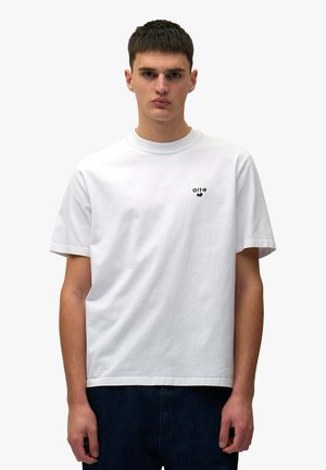 Giovane uomo che indossa una t-shirt bianca a maniche corte con un piccolo logo nero "arte" e jeans blu, in piedi contro uno sfondo grigio chiaro.