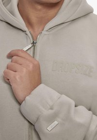 Light beige zip-up hoodie made from soft fabric, features a logo print on the chest and tag on the sleeve, with a drawstring hood.