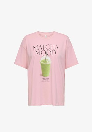Jasnoróżowy t-shirt z napisem „MATCHA MOOD” nad zielonym napojem matcha w plastikowym kubku oraz „GREEN IS THE NEW BLACK” poniżej.