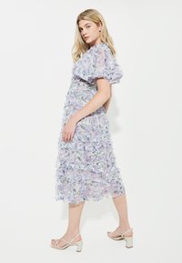 Lavendelfarbenes Blumenkleid aus transparenter Stoff, mit rüschierten Lagen und einer Länge bis unter das Knie. Kombiniert mit hellen, hohen Sandalen.