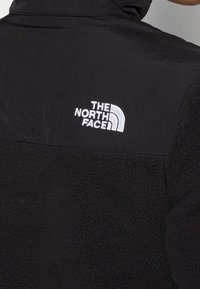 Chaqueta de forro polar negra con una sección superior suave, que presenta un logo bordado blanco en la parte posterior. Textura suave y con un cuello alto.