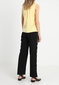 Top amarillo sin mangas con una textura suave, combinado con pantalones de pierna ancha negros que presentan un detalle de botón lateral. Unos zapatos negros sin hebillas completan el conjunto.