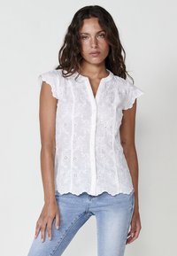 Blouse blanche brodée avec des manches tombantes festonnées, un bouton sur le devant et des motifs floraux. Tissu léger avec une finition texturée.