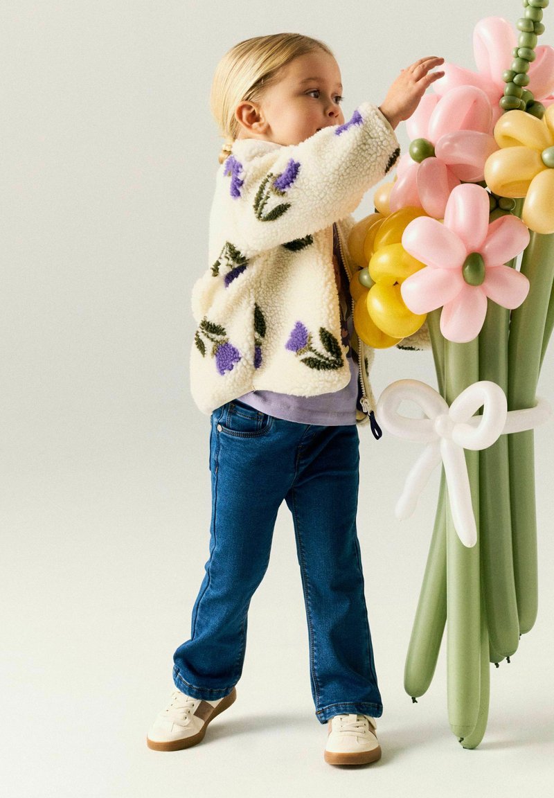 Enfant en veste fleurie tendant la main vers un grand bouquet de ballons en forme de fleurs attaché avec un nœud en ballon.