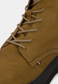 Botte haute en daim marron avec tige texturée, lacets beige, et un petit accent rouge, blanc et bleu près de la semelle. Semelle extérieure en caoutchouc noir.
