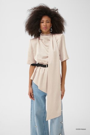 Kvinde med voluminøst krøllet hår iført en sart lyserød asymmetrisk satin-top med sort bælte og lyseblå viddeben-jeans med knapper.