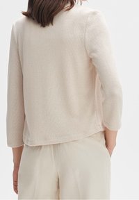 Jasno beżowa, teksturowana kurtka dzianinowa z rękawami trzyczwartowymi, nieco dłuższym tyłem i luźnym krojem. Dopasowana z jasnymi spodniami.