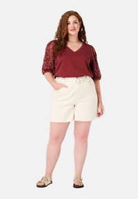 Chemise V beige à col en V avec des manches bouffantes à motifs, associée à un short crème avec une taille ceinturée. Des sandales légères complètent la tenue.
