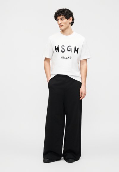 Jeune homme aux cheveux bouclés portant un t-shirt blanc MSGM Milano, un pantalon large noir et des chaussures noires, debout les mains dans les poches.