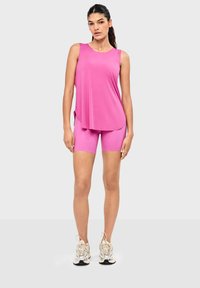 Femme debout portant un haut rose ample sans manches, un cuissard rose assorti et des baskets beiges sur un fond clair uni.