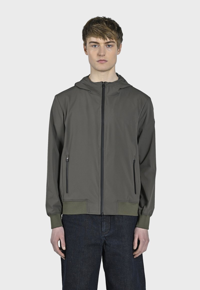 Kapuljača jakna u maslinastoj boji, sa zatvorenim prednjim džepovima, elastičnim manžetama i glatkom teksturom tkanine. Nosena preko tamnih traperica.