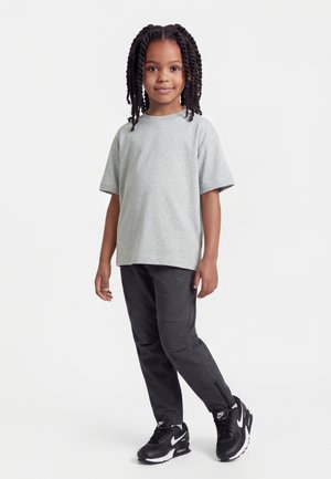 Niño con cabello trenzado que lleva una camiseta de manga corta gris claro, pantalones negros y zapatillas negras Nike, de pie contra un fondo blanco.