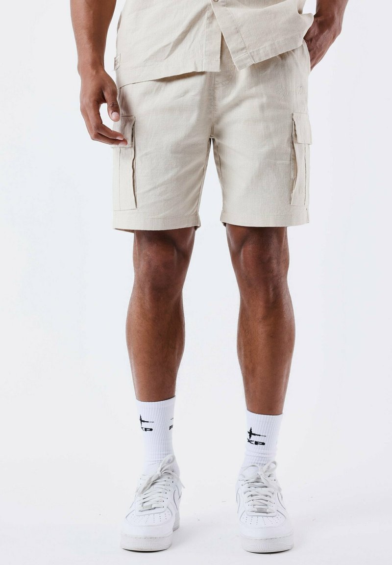 Shorts cargo beiges en tissu texturé, dotés de deux poches latérales, d'une coupe décontractée et d'une longueur au-dessus du genou. Portés avec des baskets blanches.