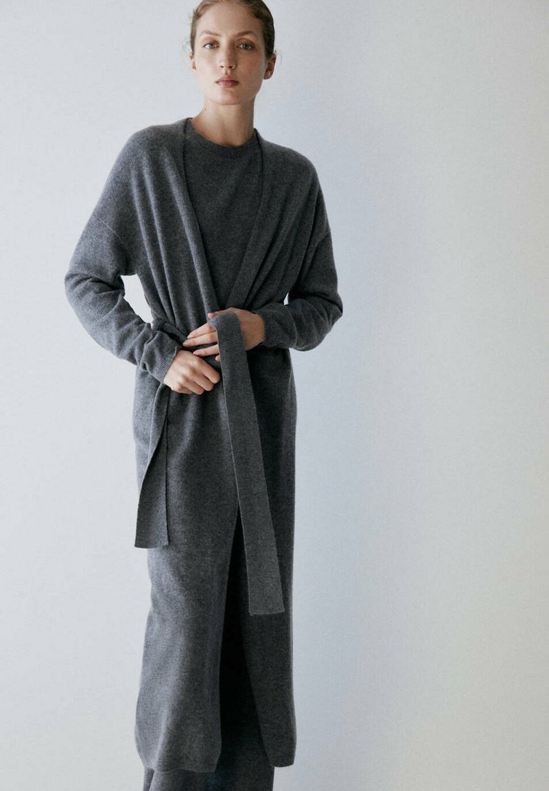 Femme portant un long cardigan gris et une robe en maille assortie, attachant la ceinture du cardigan, debout devant un fond clair uni.