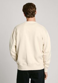 Sudadera color crema con cuello redondo, puños y dobladillo acanalados, y una textura suave; cuenta con hombros caídos para un ajuste relajado.