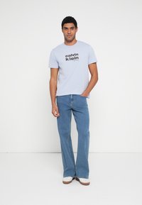 Hele sinine puuvillane T-särk musta "calvin klein" teksiga, kombineeritud hele siniste lehvikpükstega ja valgete jalanõudega, millel on beežid tallad.