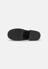 Chaussure noire à enfiler avec une semelle en caoutchouc texturé, un matériau supérieur lisse et un design minimaliste, présentant un dos ouvert bas pour un enfilage facile.