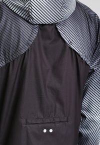 Giacca nera con cappuccio a righe grigie. Presenta una tasca con due bottoni bianchi e un tessuto morbido e leggero.