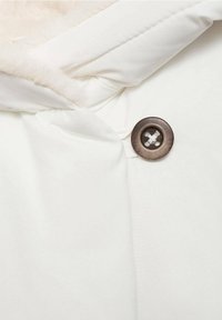 Weißer Stoff mit weichem, strukturiertem Innenfutter. Ein runder, dunkelbrauner Knopf mit gesticktem Muster verschließt das Kleidungsstück.