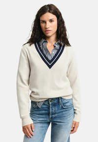 Cremfarbener V-Ausschnitt-Pullover mit marineblauen Akzenten, über einem gestreiften Hemd getragen. Weiche Textur, lockere Passform und gerippte Bündchen, kombiniert mit blauen Jeans.