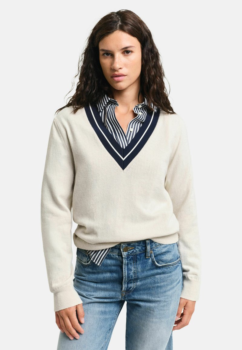 Cremfarbener V-Ausschnitt-Pullover mit marineblauen Akzenten, über einem gestreiften Hemd getragen. Weiche Textur, lockere Passform und gerippte Bündchen, kombiniert mit blauen Jeans.