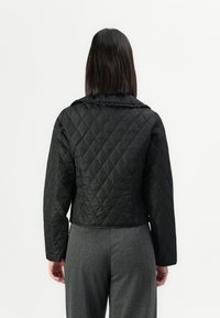Femme aux cheveux raides et foncés portant une veste matelassée noire et un pantalon gris, se tenant de dos face à l'appareil photo sur fond blanc.