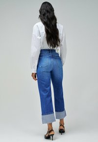 Jeans de mezclilla azul de tiro alto con un corte de pierna ancha y puños en contraste de azul claro. Combinados con una camisa blanca de botones y tacones negros.