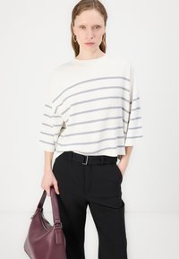 Pulover tricotat cu dungi în crem cu dungi gri, mâneci largi; asociat cu pantaloni negri și o geantă burgundă.