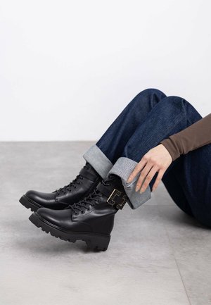 Botas negras de cuero hasta el tobillo con suela gruesa, diseño con cordones y detalle de hebilla dorada. Combinadas con jeans de mezclilla azul con puños.