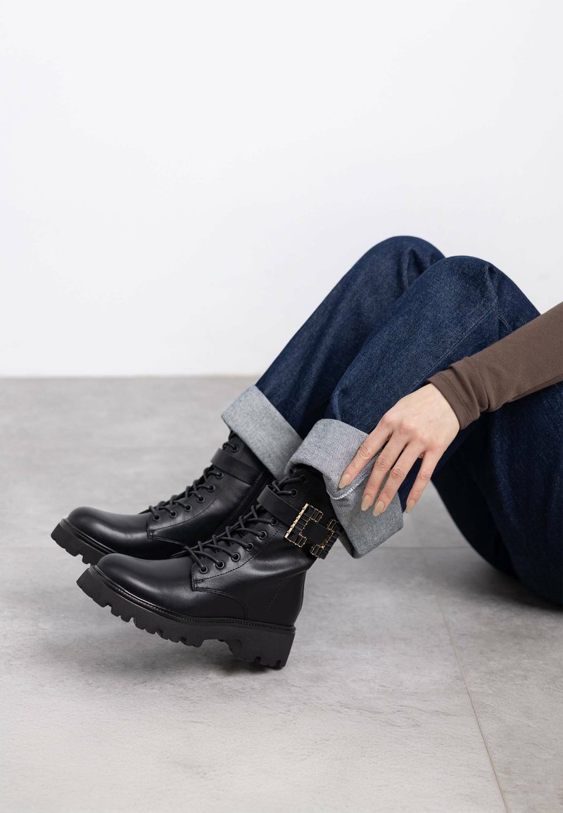 Botas negras de cuero hasta el tobillo con suela gruesa, diseño con cordones y detalle de hebilla dorada. Combinadas con jeans de mezclilla azul con puños.