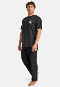 Black cotton t-shirt with short sleeves and a round neckline, featuring a circular graphic patch on the chest. Paired with loose black pants.