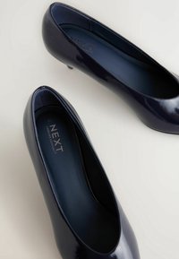 Escarpins en cuir vernissé bleu marine avec une finition lisse, bout rond et talon bas. L'intérieur comprend une semelle rembourrée et un logo de marque.